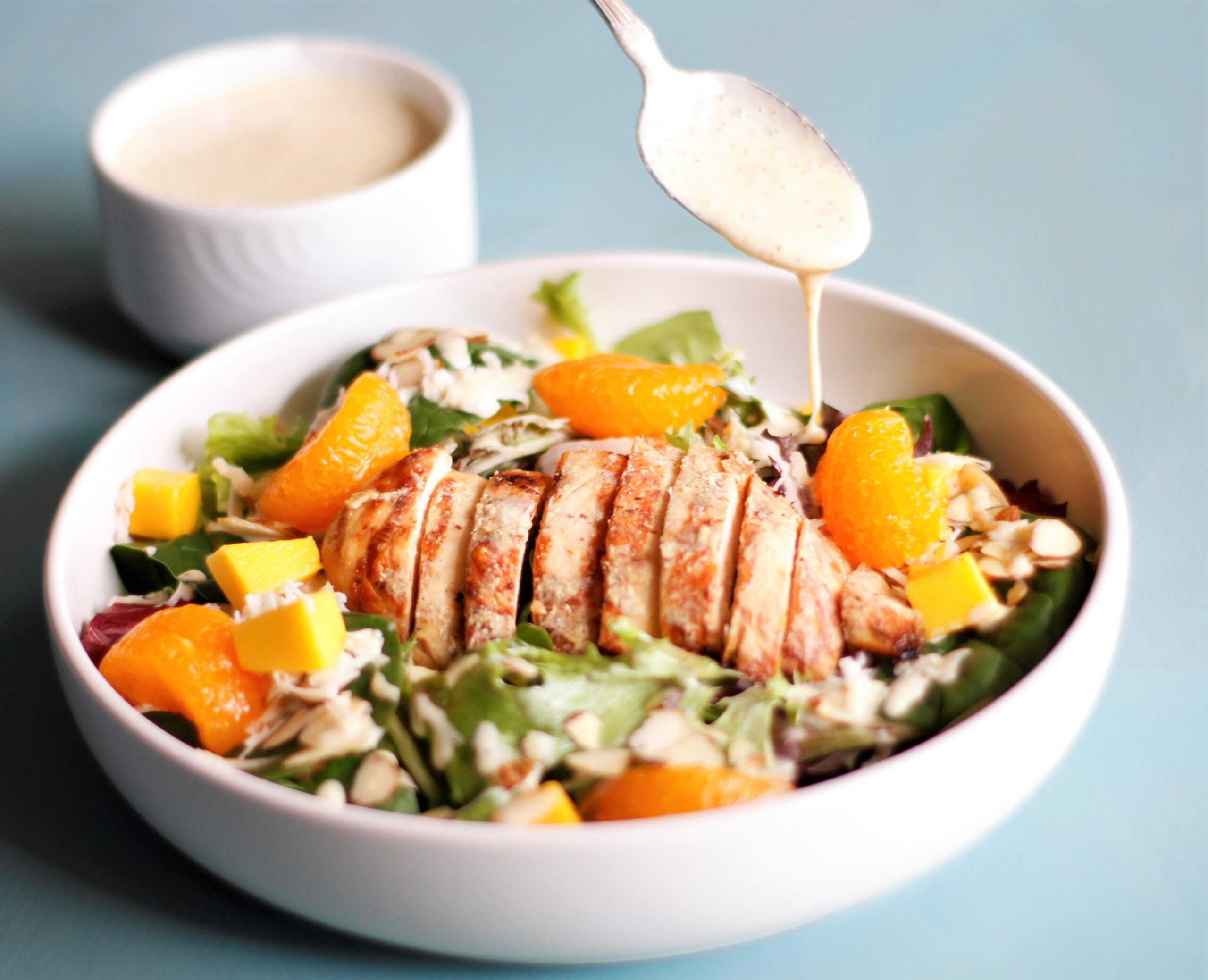 A salad with grilled chicken, mandarin oranges, almonds and flaked coconut with a spoonful of honey mustard dressing being drizzled over top.