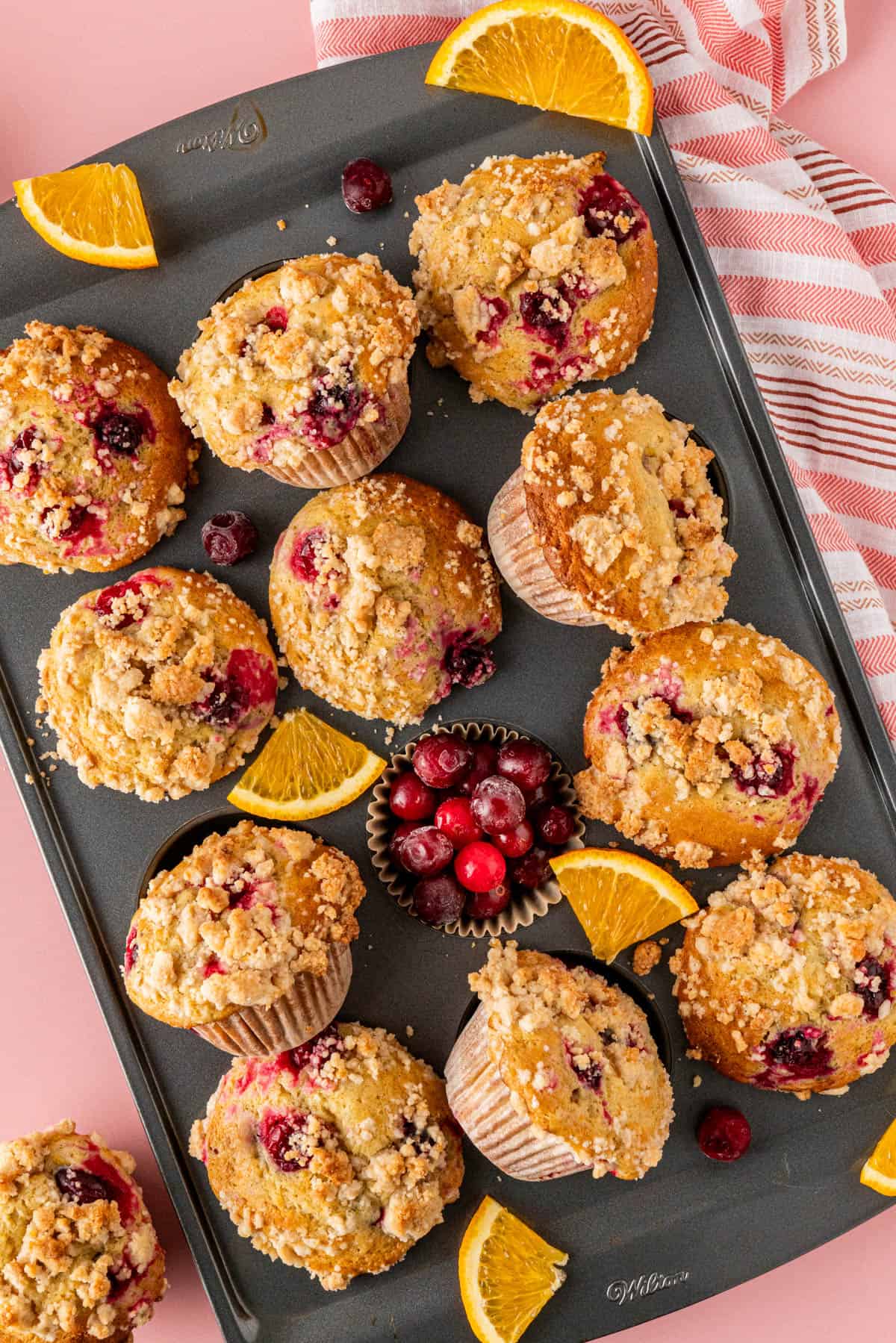 A 12-piece muffin pan filled with 11 pieces of golden baked Cranberry Orange Muffin and a brown liner filled cranberries.  