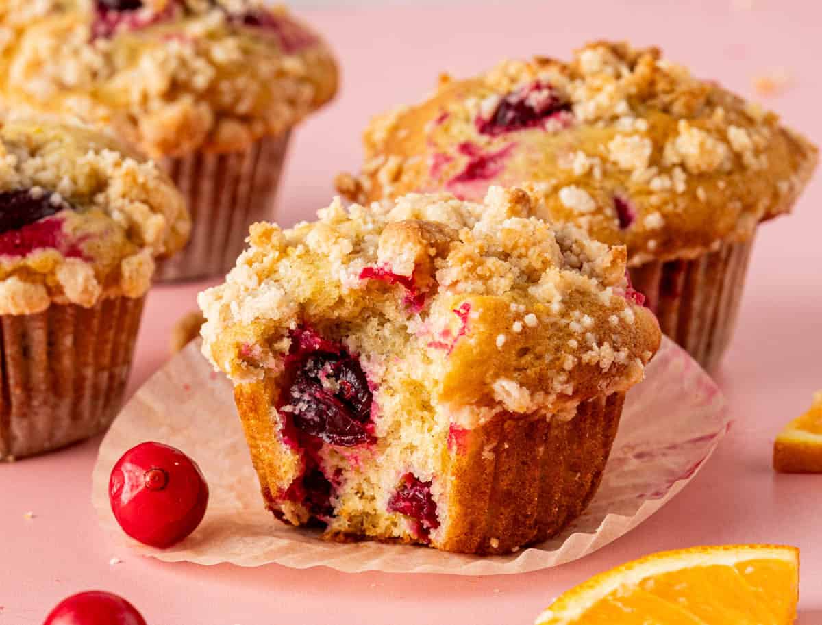 Cranberry orange muffins with streusel topping. One bitten to show interior. Fresh cranberries and orange slices on the sides. 