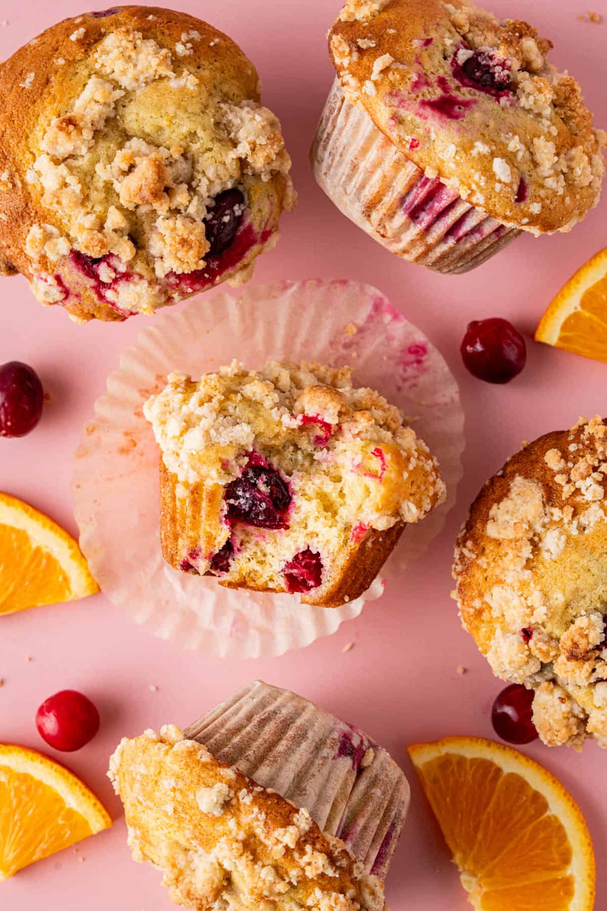 Cranberry orange muffins with crumb topping arranged on pink surface; one bitten to show moist interior. Surrounded by fresh cranberries and orange slices.