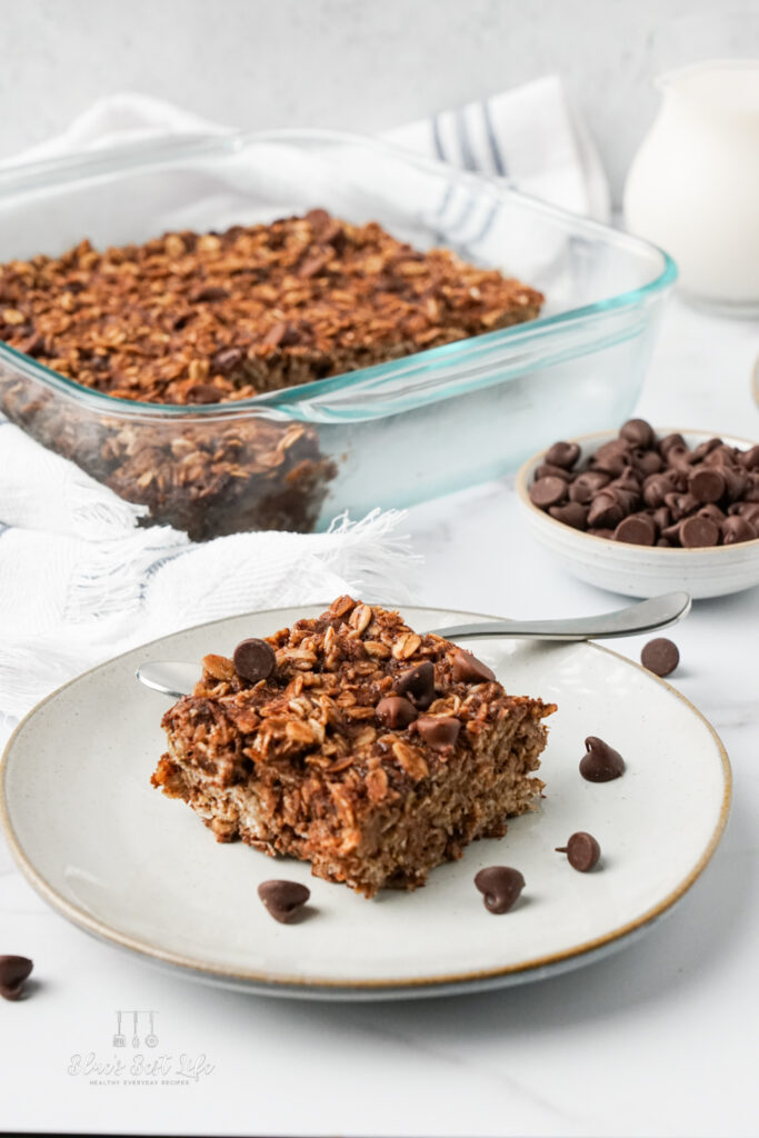 A piece of chocolate baked oatmeal on a plate with the serving dish and extra chocolate chips.  