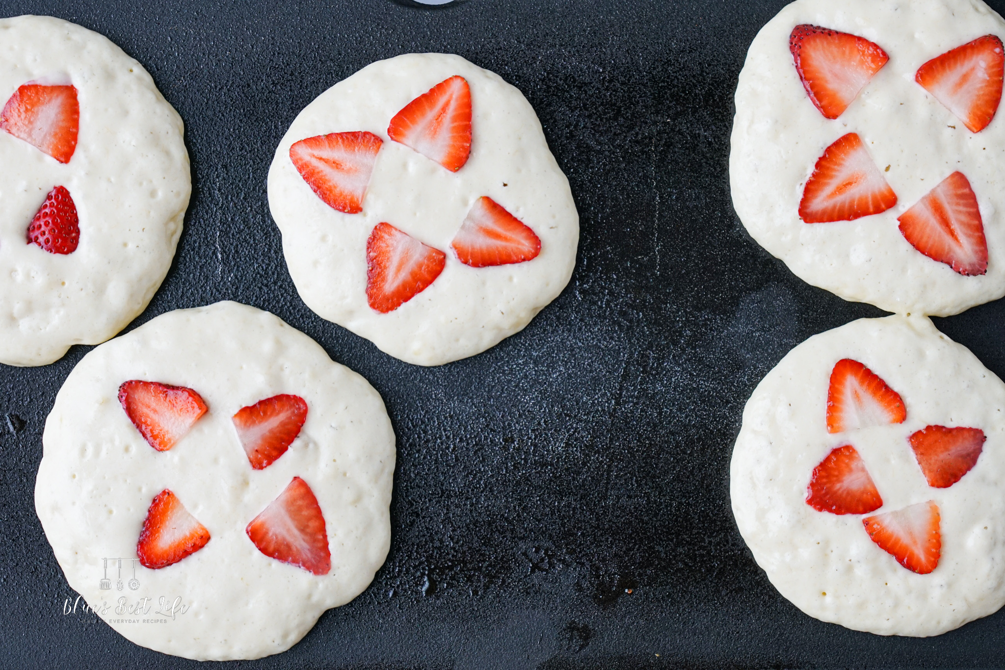 Five uncooked strawberry-topped pancakes on a griddle, decorated with quartered strawberries arranged in a star-like pattern.