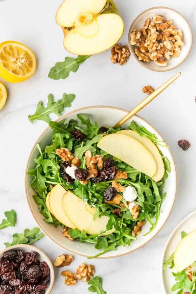 A bowl of arugula salad with dried cherries, walnuts, apples and goat cheese. 