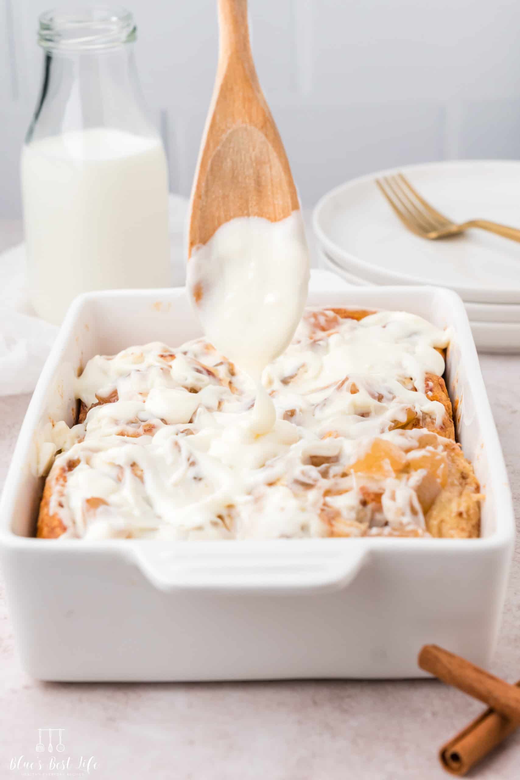Close-up of cinnamon rolls in white dish with cream cheese frosting being drizzled from wooden spoon; background includes milk bottle, stacked plates, and gold forks.