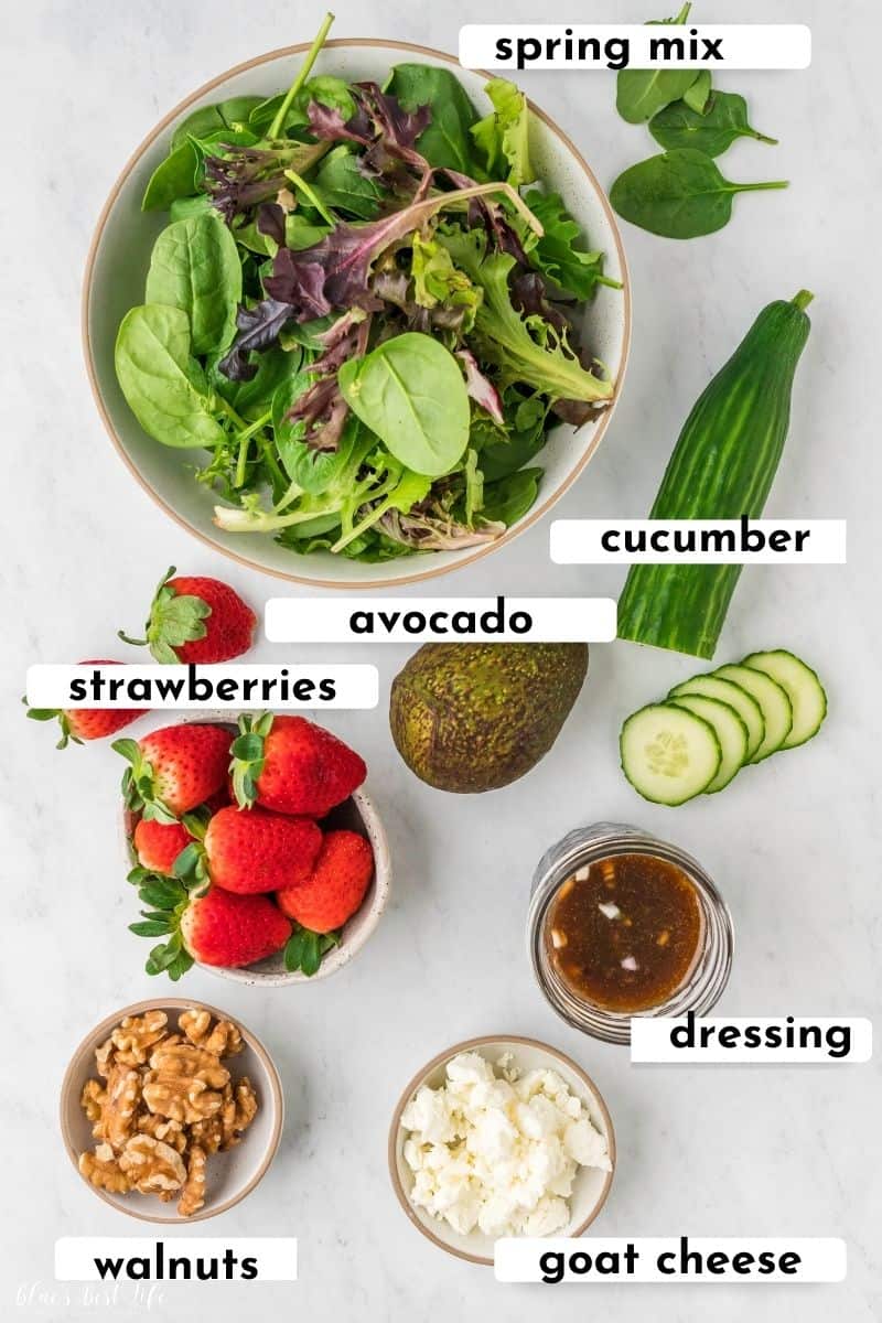 The ingredients to make strawberry walnut salad: A bowl of spring mix, cucumber, avocado, strawberries, dressing, walnuts, goat cheese. 