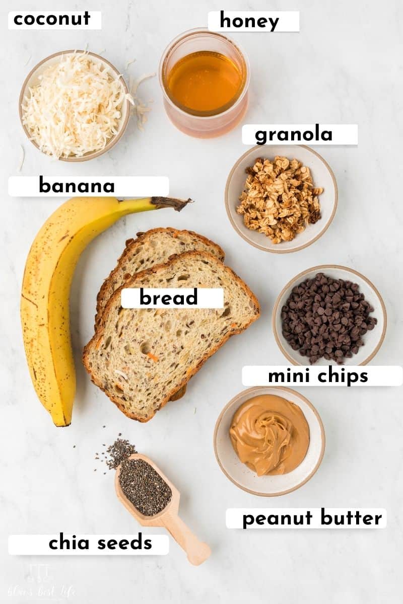 Flat lay of the ingredients to make peanut butter banana toast. such as: coconut, honey. banana. bread, mini chocolate chip, granola, peanut butter, and chia seeds.