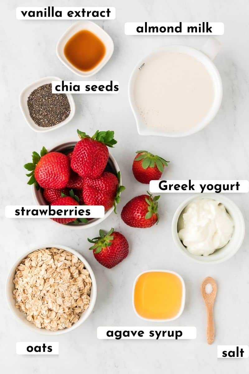 Flat lay of the ingredients for strawberry shortcake overnight oats such as: vanilla extract, almond milk, chia seeds, strawberries, Greek yogurt, oats, agave syrup, salt.