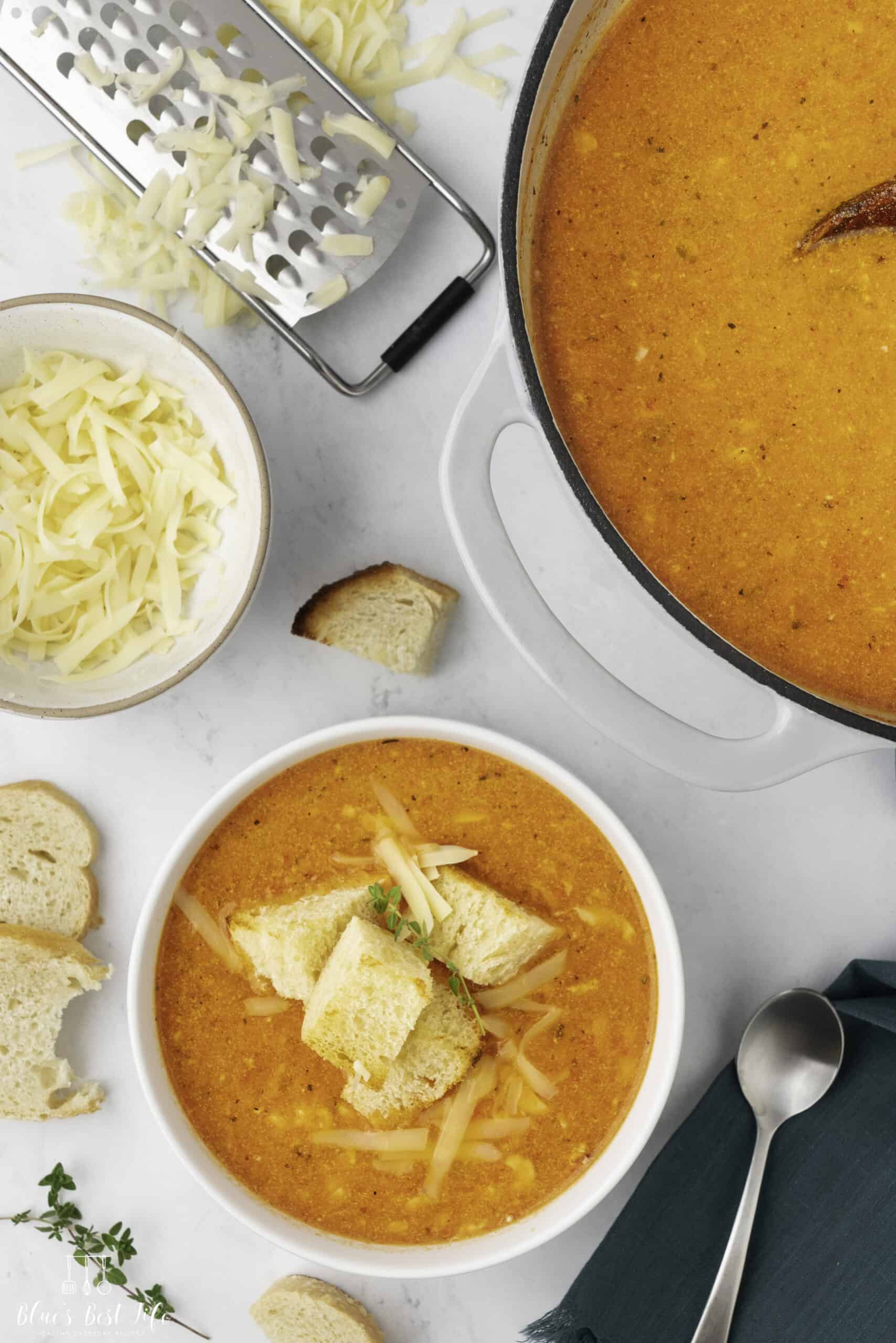 A bowl of roasted red pepper gouda soup, a bowl filled with shredded cheese and a pot of red pepper soup on the sides.