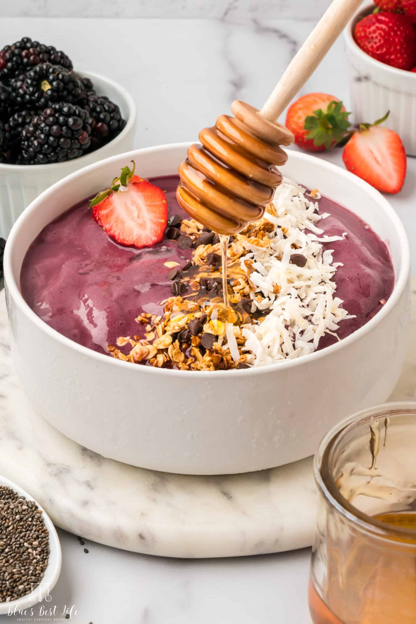 Drizzling the smoothie bowl with honey using a honey wand; on the sides separate bowls of raspberries and strawberries can be seen.
