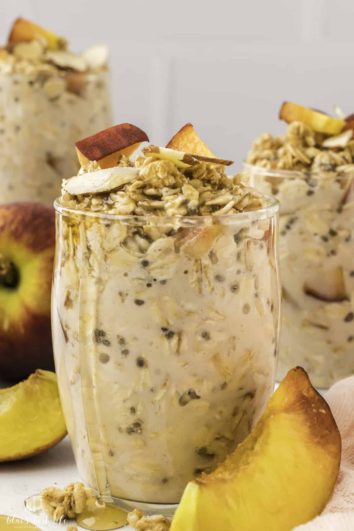 A glass jar of overnight oats with peaches and honey drizzling. Two more servings is blurry but still visible in the background. 