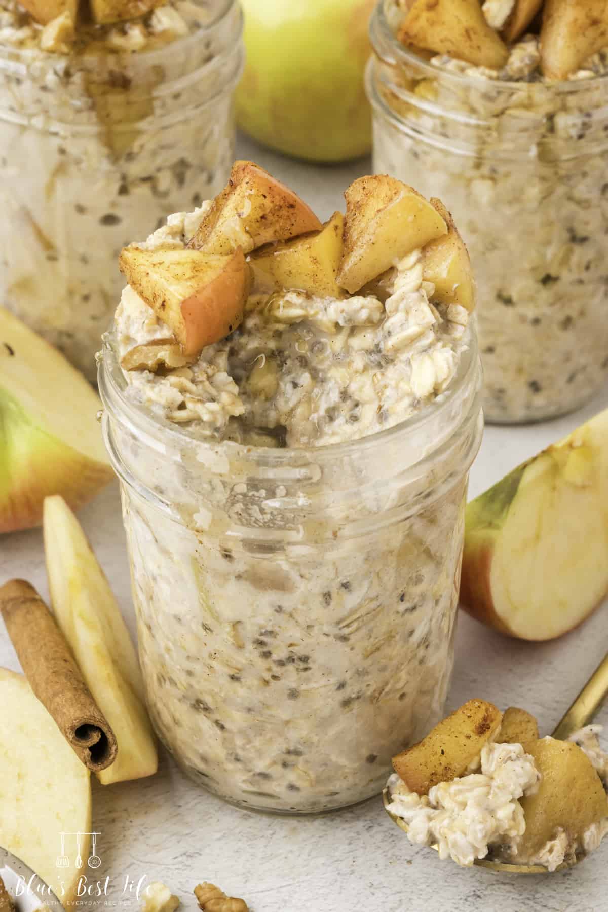 Three Glass jars of overnight oats mixed with chia seeds and topped with cinnamon apples; surrounded by fresh apple slices and a cinnamon stick.