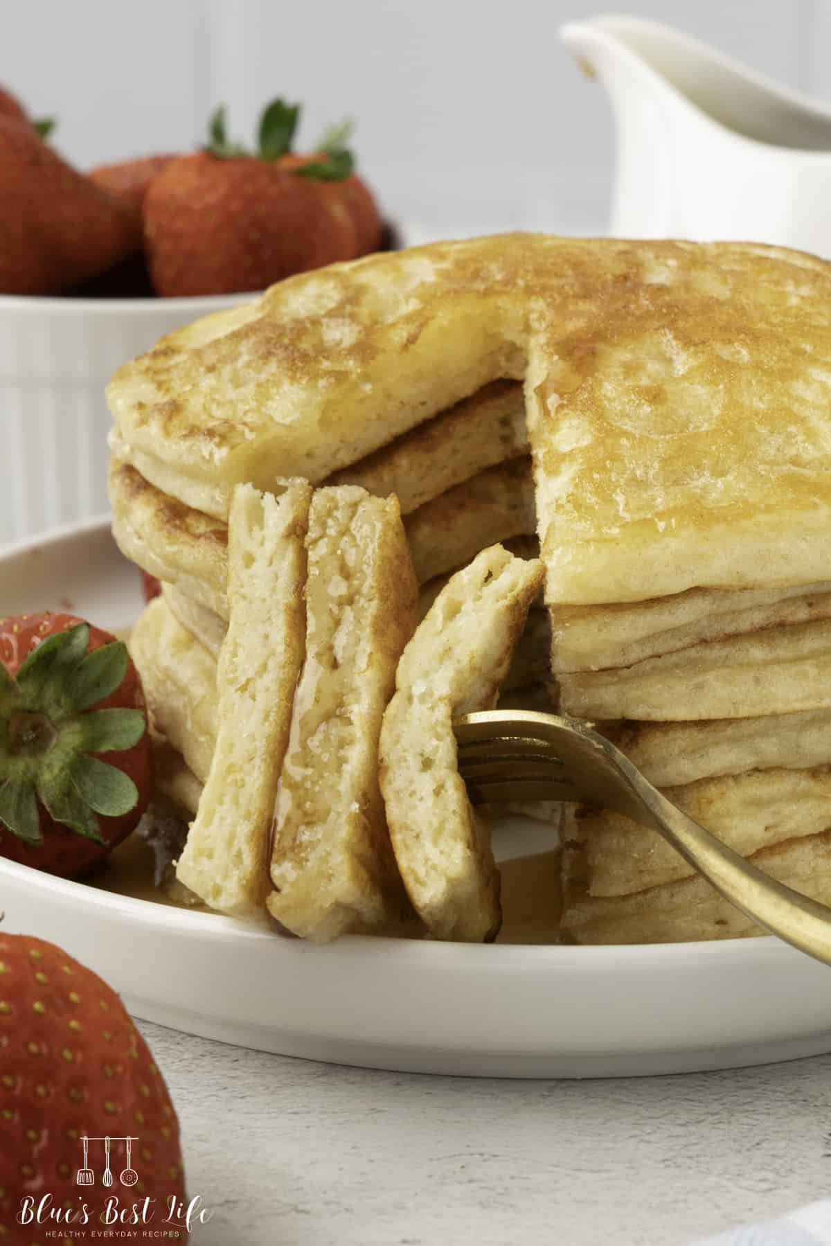 Stack of fluffy pancakes with syrup on white plate; fork lifts a cut portion to show airy texture. Background includes bowl of strawberries and ceramic pitcher.