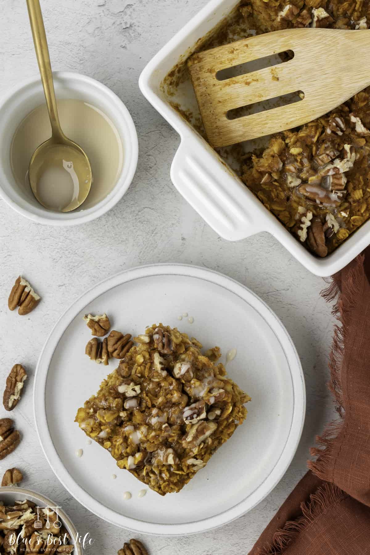 A piece of the pumpkin oatmeal with vanilla glaze served on a white ceramic plate. glaze sitting on a ceramic bowl on the side and the baking dish filled with th e pie.