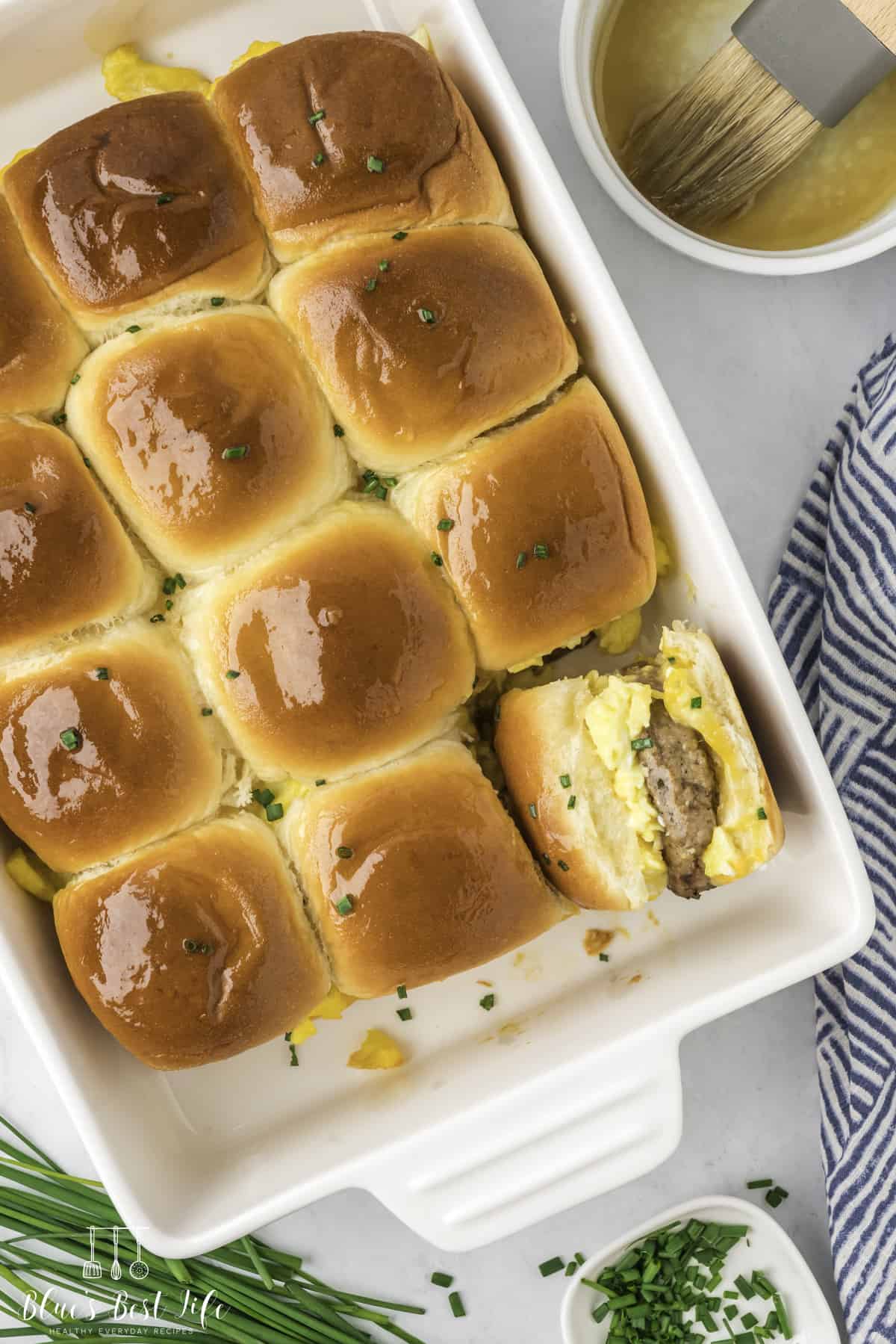 Breakfast sliders in baking dish made with golden dinner rolls; one removed to reveal scrambled eggs, cheese, and sausage patty filling. Garnished with chives, served beside melted butter and striped cloth.