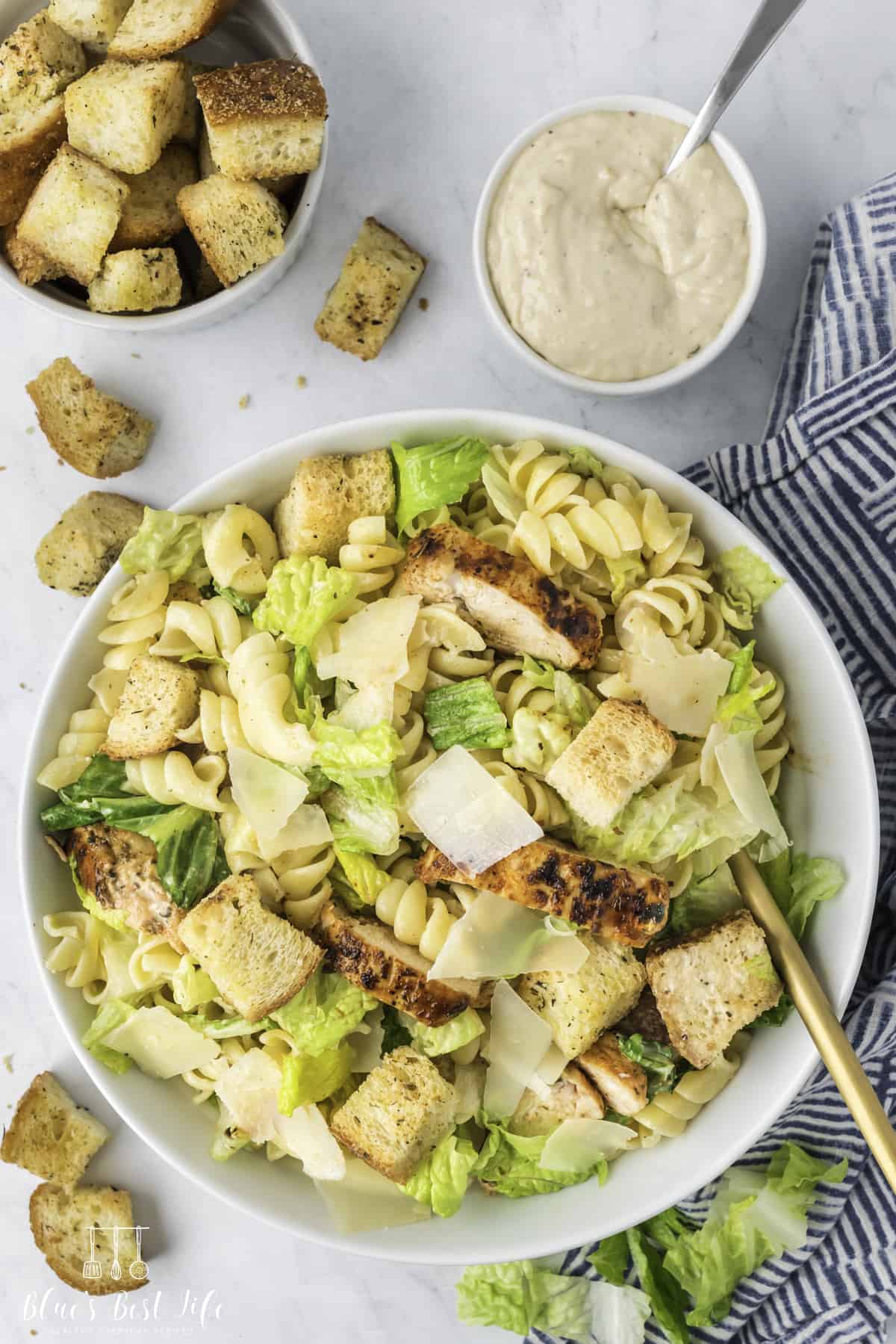 Top shot of a bowl with the chicken Caesar pasta salad, a small bowl of Caesar dressing and a bowl of croutons on the side. 