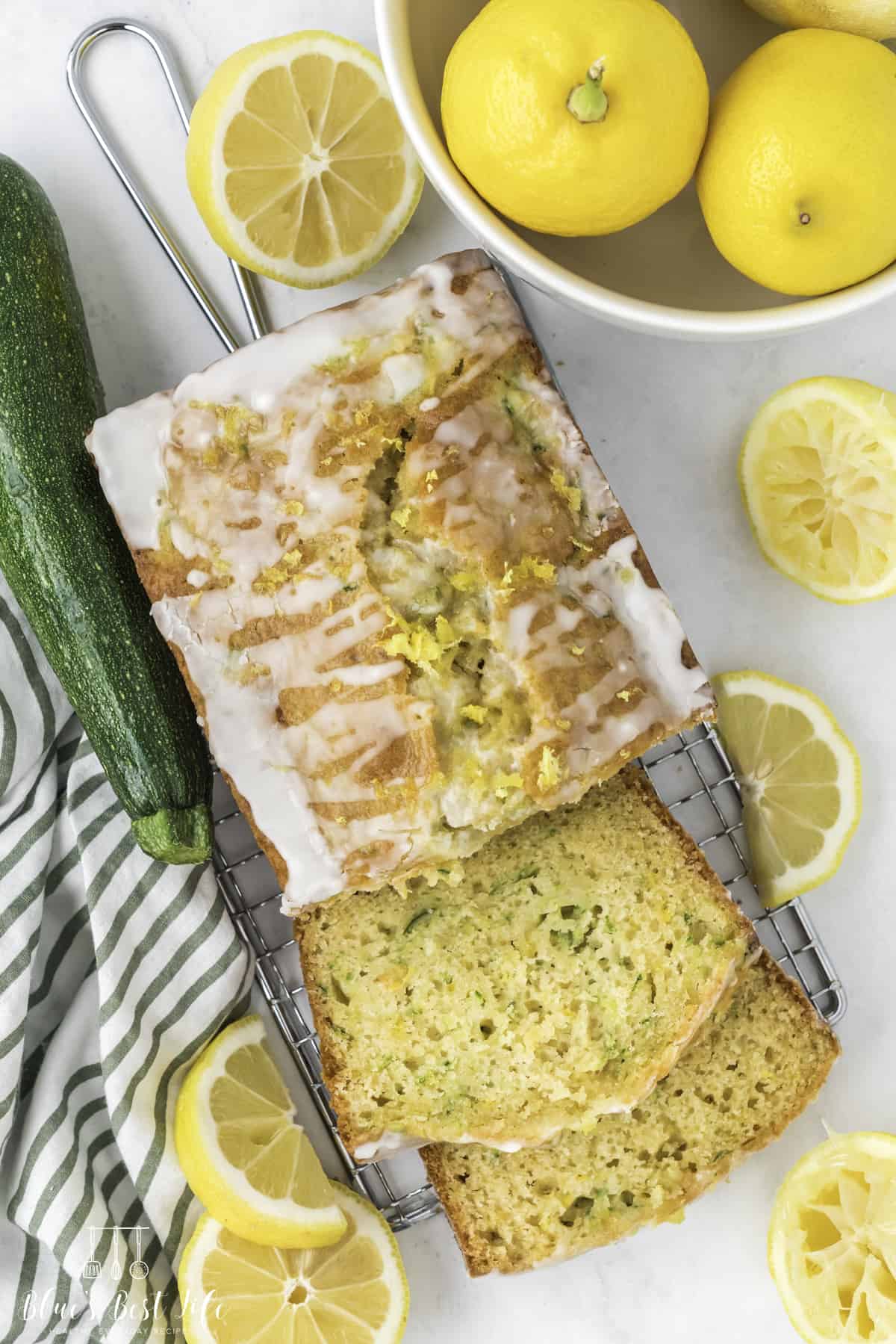 The lemon zucchini bread on a rack, two slices can be seen. zucchini and lemon is also on the sides.