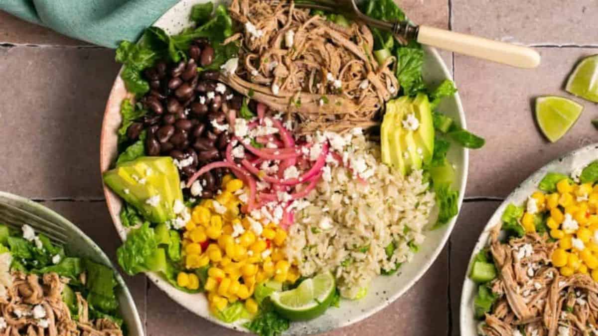 a burrito bowl with pork carnitas, avocado slices, red onion, beans, corn, rice, and salad