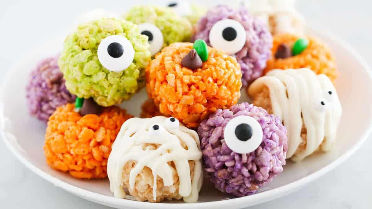 on a plate rice krispie treats balls in orange pumpkin designs, white mummy, and purple and green balls with googly eye each