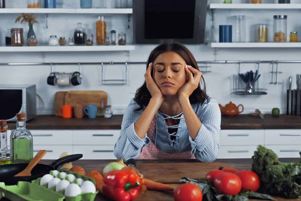 Tired woman eyes closed with ingredients around her in the kitchen.