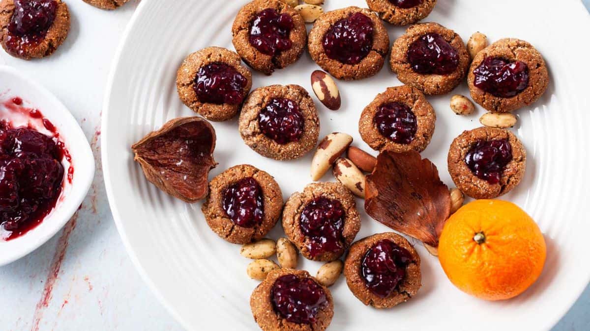 Thumbprint cookies in a white plate with orange, almond nuts, and unsweetened jam on the side.