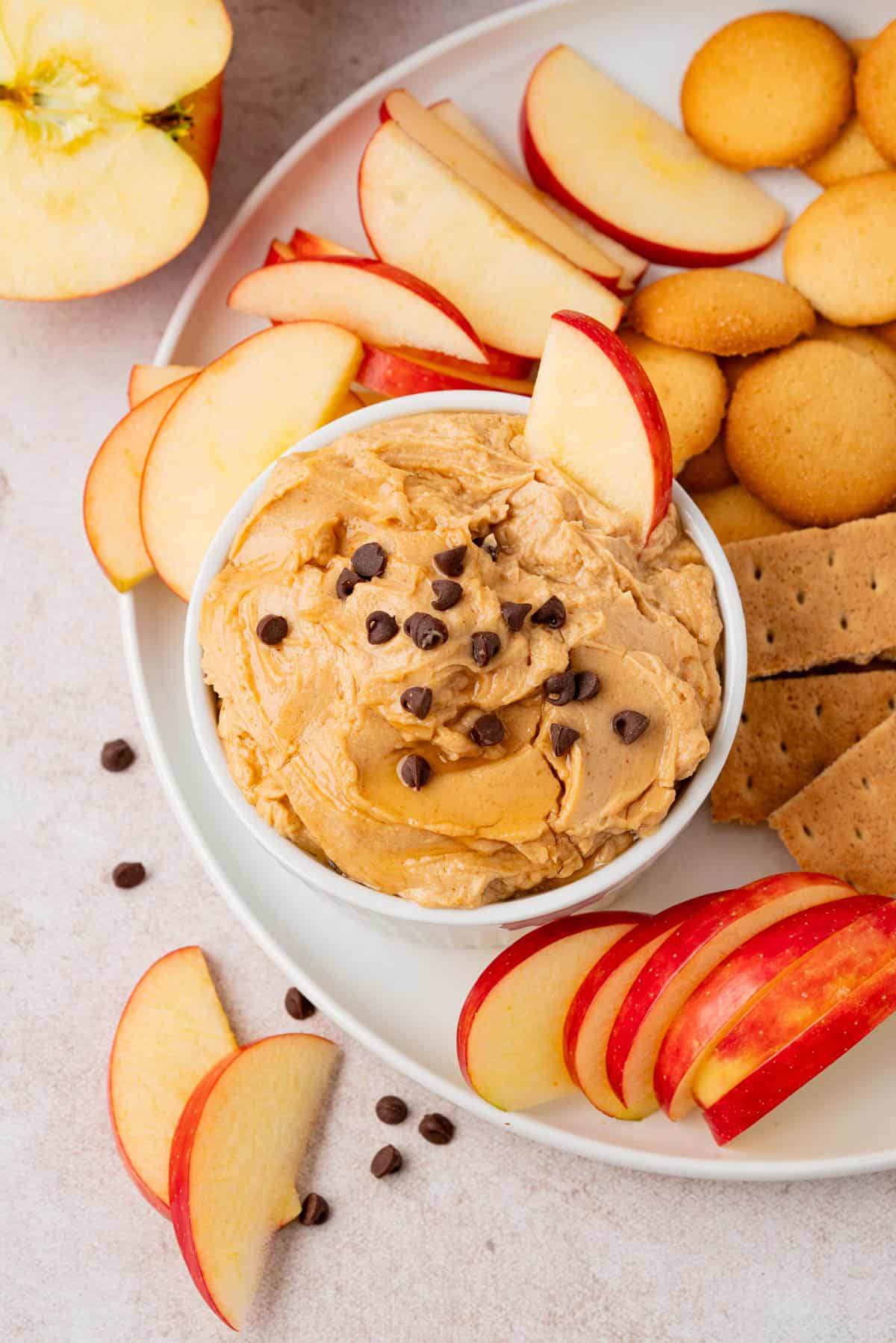 Peanut Butter Dip With Greek Yogurt in a white small bowl with chocolate chips on top and a sliced apple dip on it. The bowl is on a plate with different dippers like sliced apples and graham crackers.