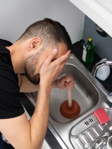 Man covering his face while trying to unclog the kitchen sink