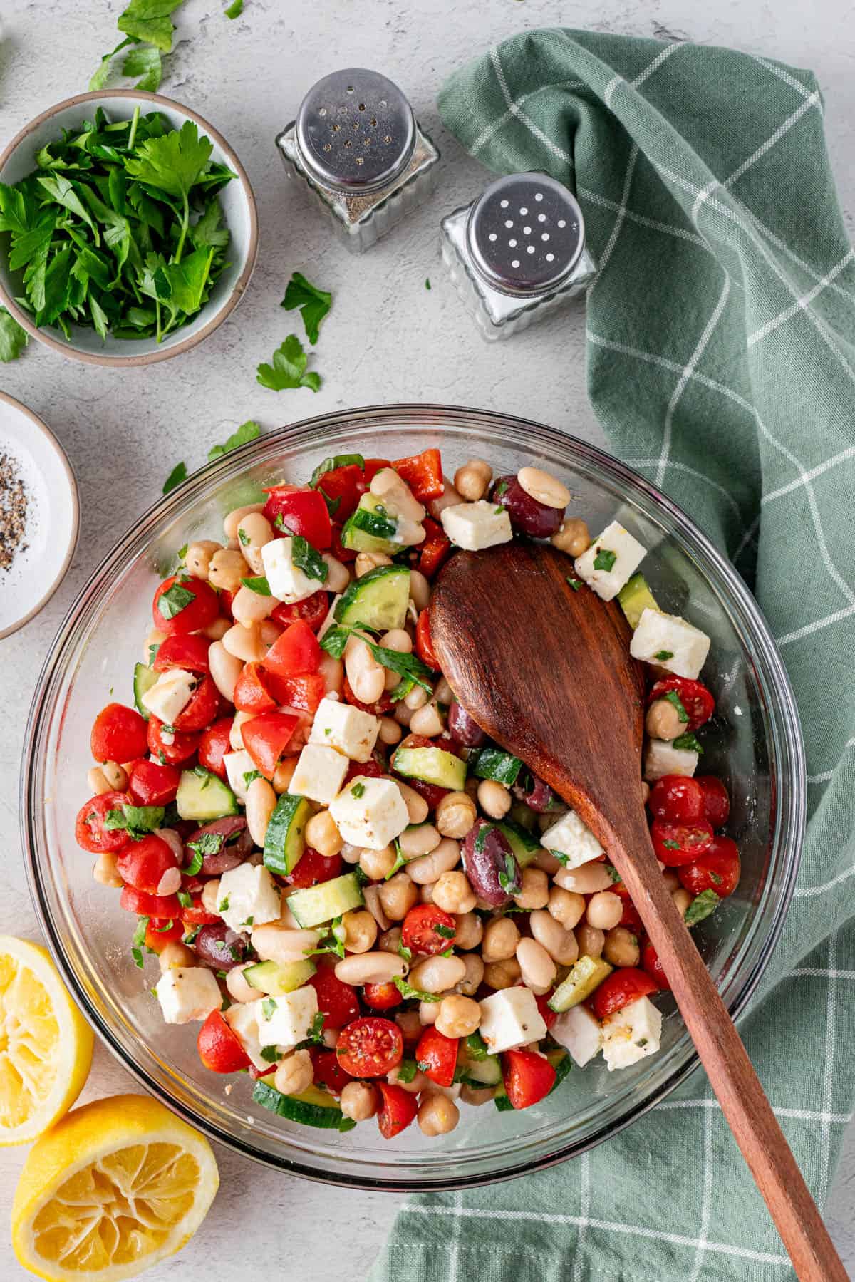 A top shot of Greek-Inspired Mediterranean Bean Salad in a clear serving bowl with a wooden spoon, salt and pepper shakers on the upper center part of the photo and some green plaid cloth. 