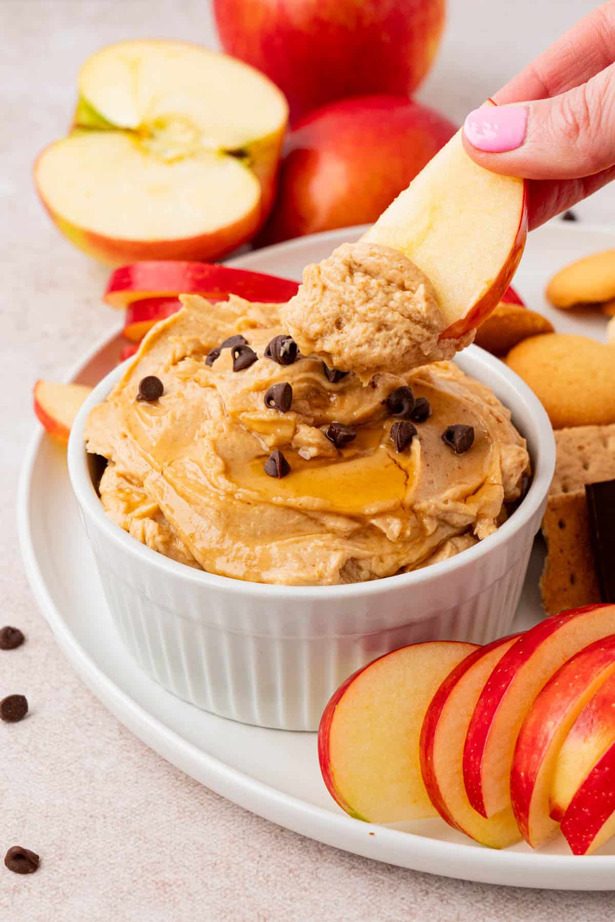 A hand holding a sliced apple dip in a small bowl of Peanut Butter Dip With Greek Yogurt with sliced apples on the background