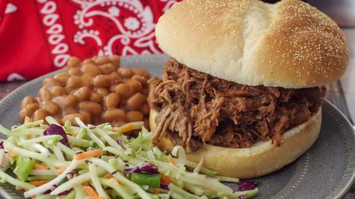 A serving of slow cooker beef on a bun with beans and veggies on a plate. 