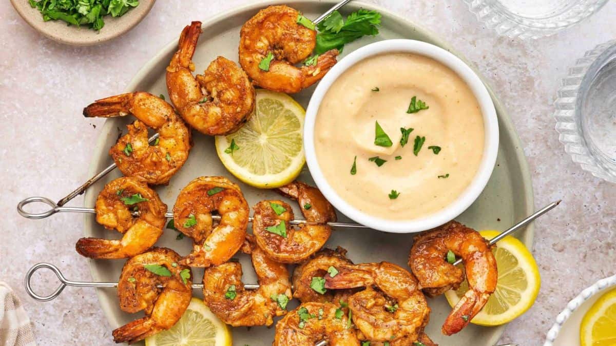 A few pieces of Oven Shrimp Skewers With Old Bay Seasoning served on a plate with a dip and a few slices of lemon. 