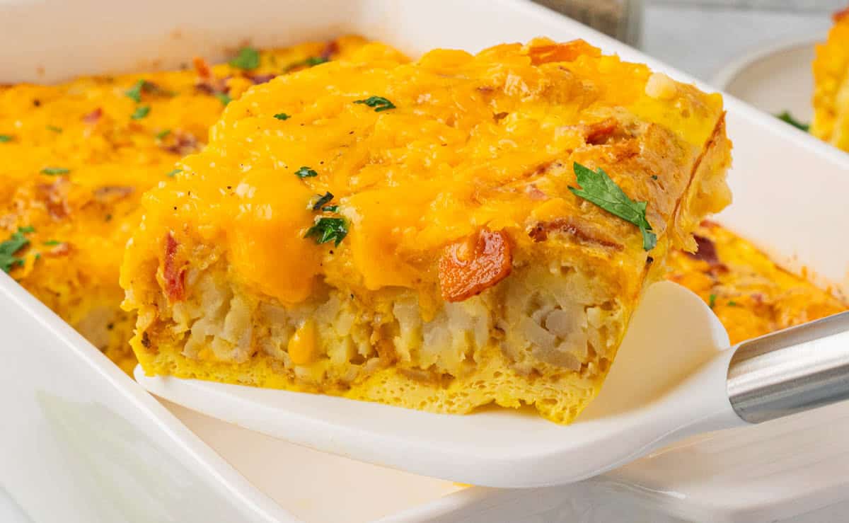 Golden Tater Tot breakfast casserole being lifted from white dish, filled with shredded potatoes, eggs, cheddar cheese, and bits of sausage or vegetables; topped with crispy layer and parsley garnish.