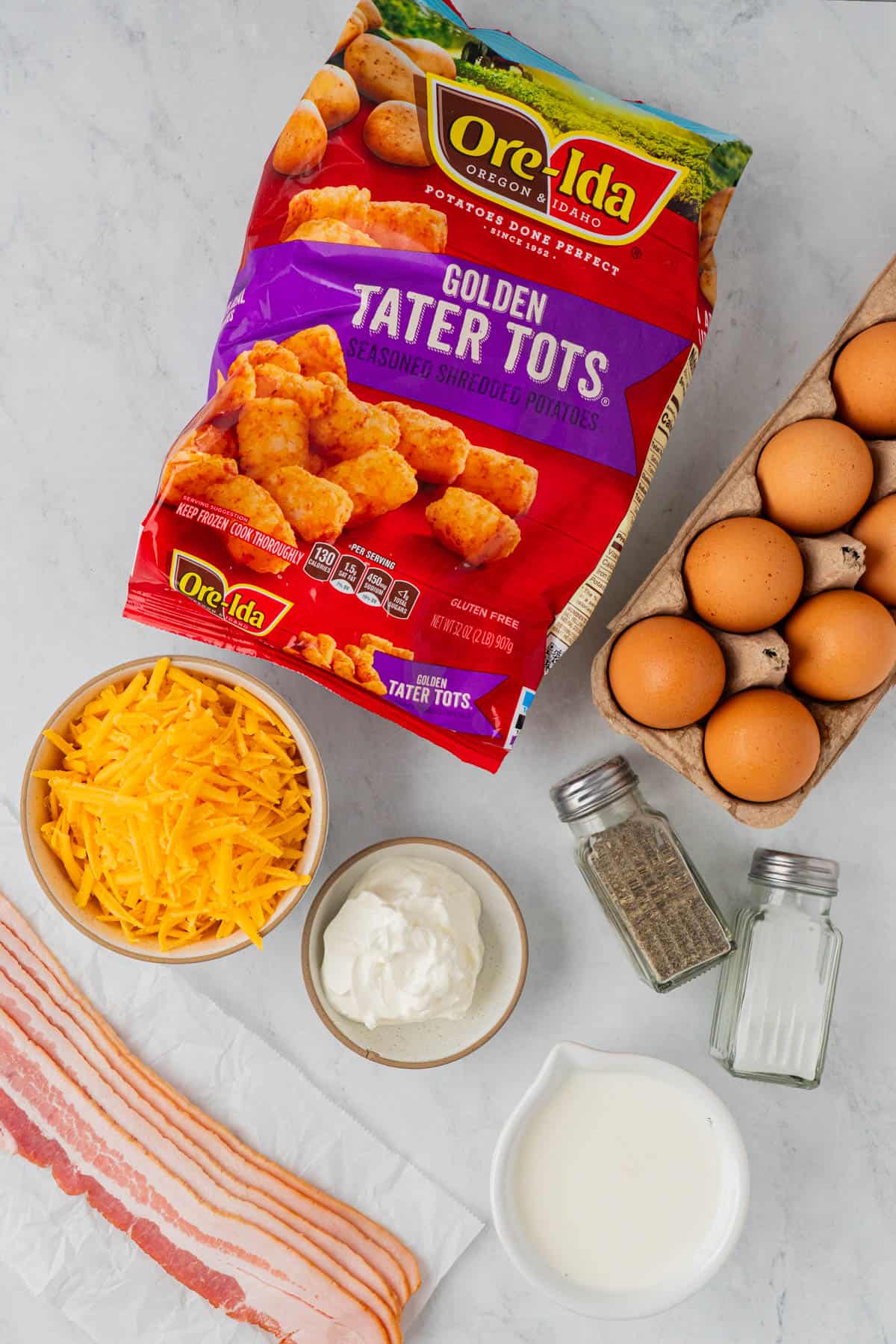 Flat lay of ingredients for the Tater Tot Breakfast Casserole With Bacon: A full bag of tater tots, eggs, cheese, sour cream, & half & half., salt & pepper, and strips of bacon. 