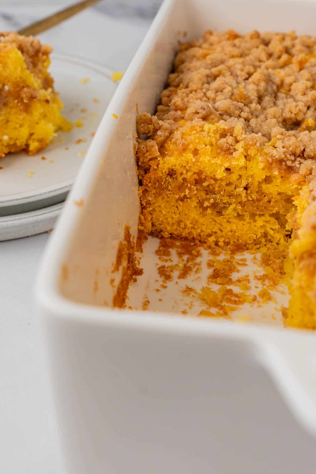 Coffee cake Mix cake in white dish with golden-orange hue and streusel topping; one slice removed to reveal interior. A plated slice sits in the background. 