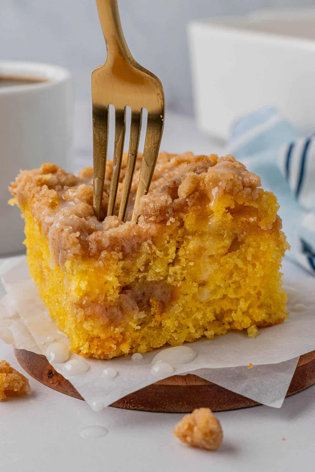 Close-up of moist coffee cake with crumbly streusel topping and light glaze; gold fork inserted into slice. Background includes coffee cup and blue-striped cloth.