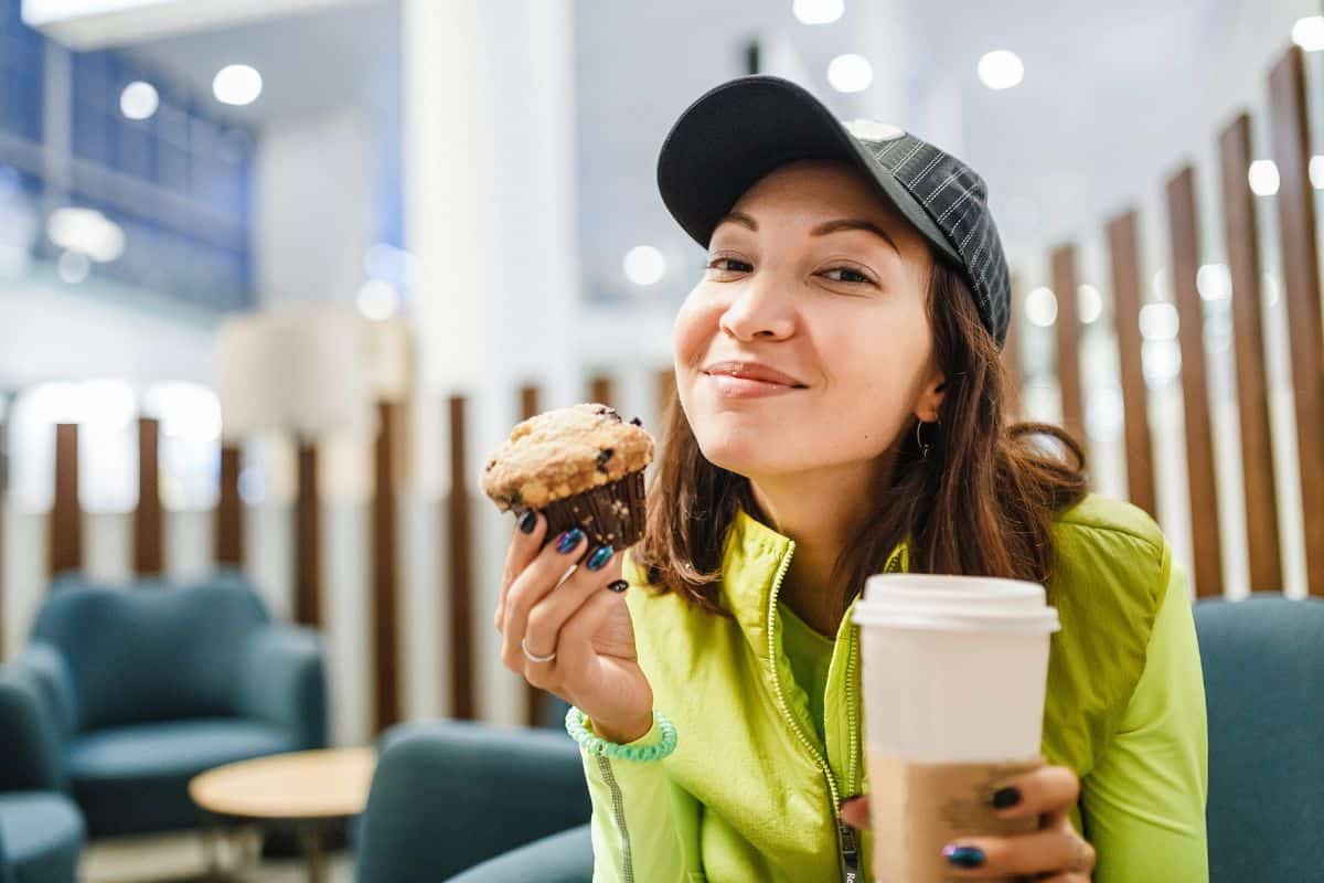 Young brunette woman drinking coffee and eating a delicious muffin cake in modern airport cafe