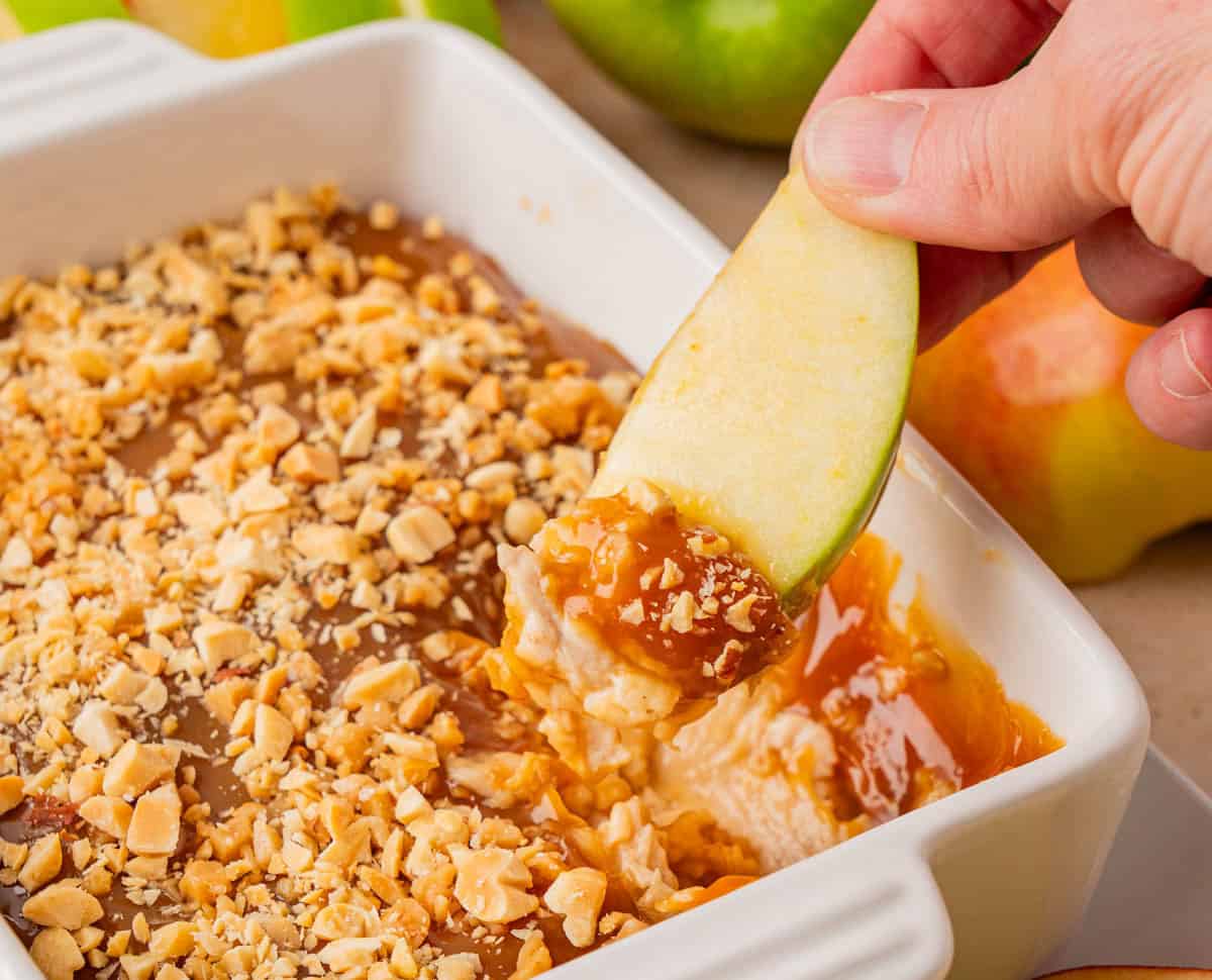 A mouthwatering caramel apple cream cheese dish and a hand holding a sliced green apple dipping on it.