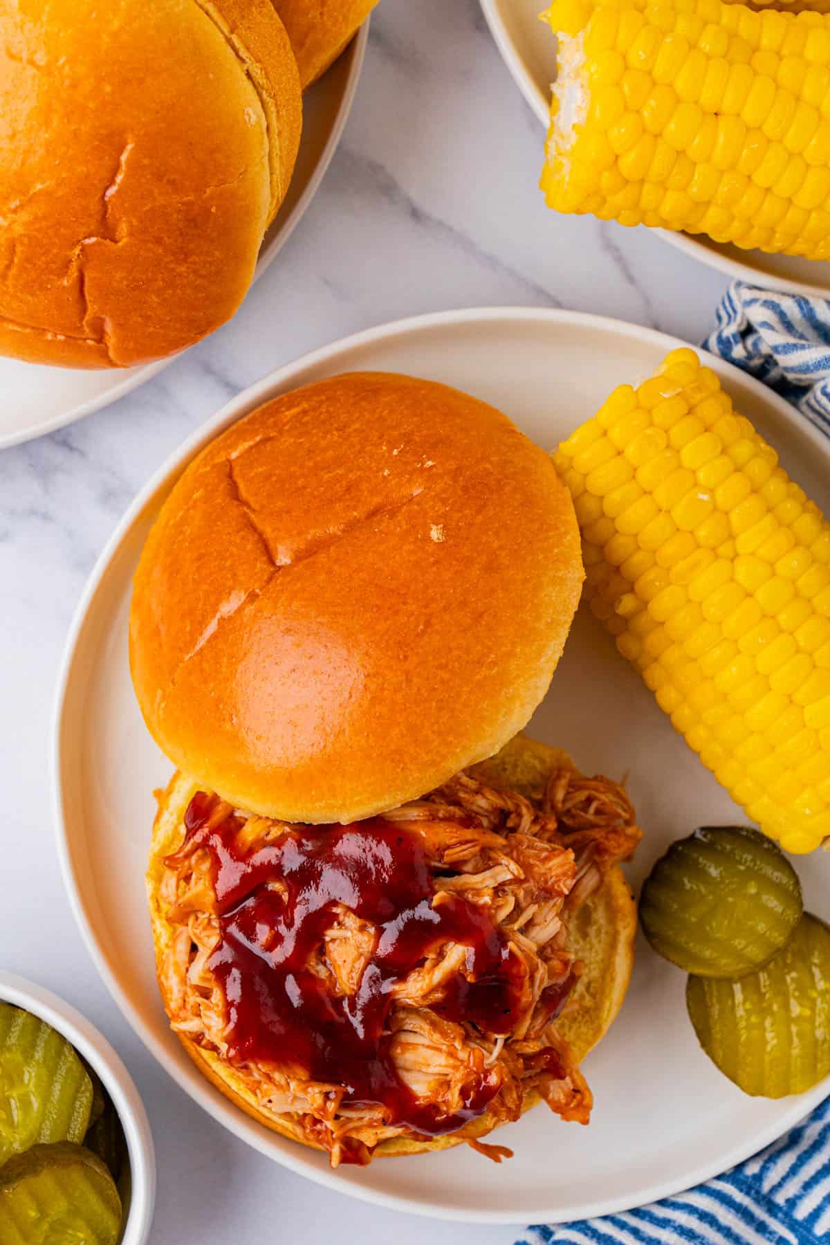 Crockpot BBQ Pulled Chicken Sandwich served in a plate with pickles and a cob of corn. 