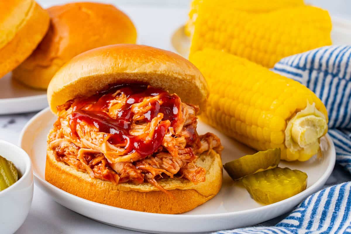 Crockpot BBQ Chicken Pulled Sandwich served on a plate with pickles and corn on cob. 