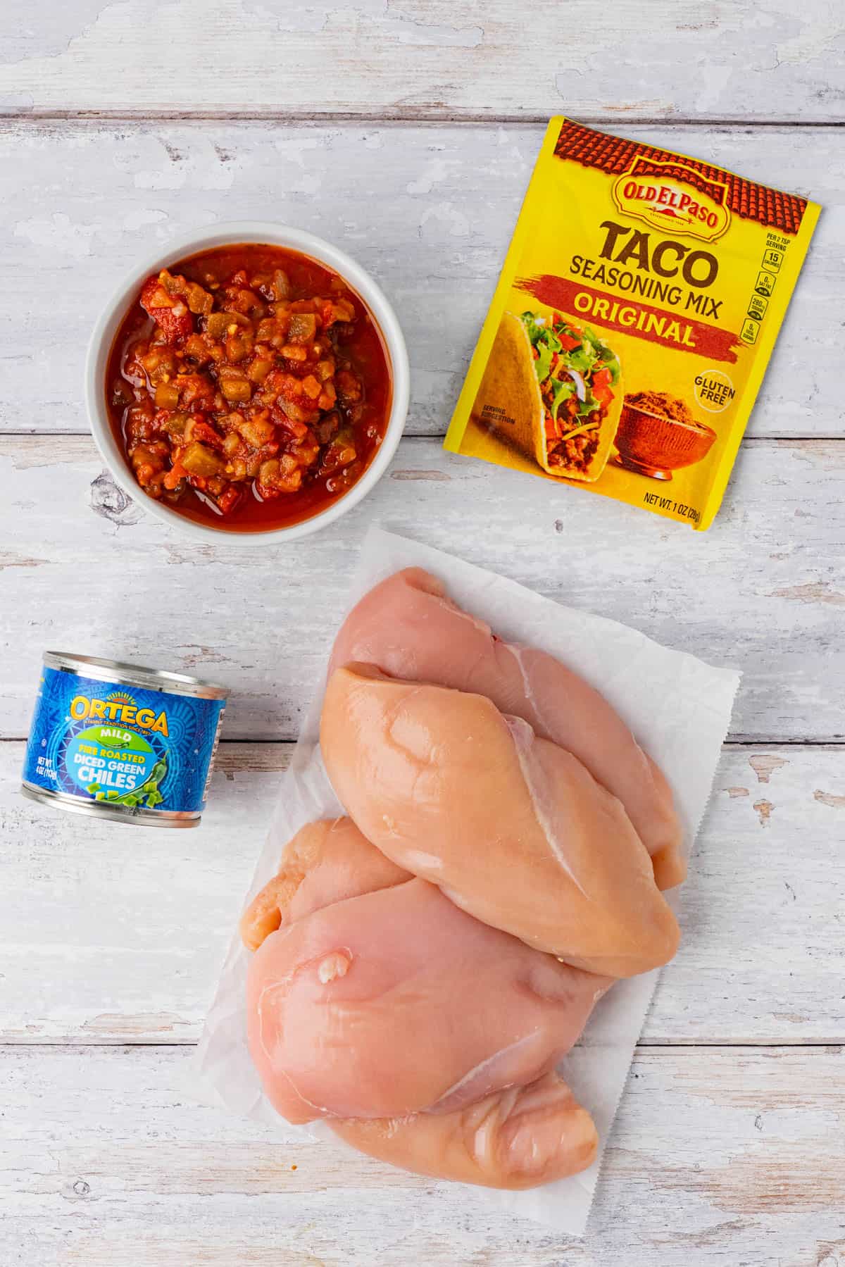 Ingredients for Crockpot Taco Chicken: salsa in a bowl, taco seasoning mix, a can of diced green chiles, chicken breasts.