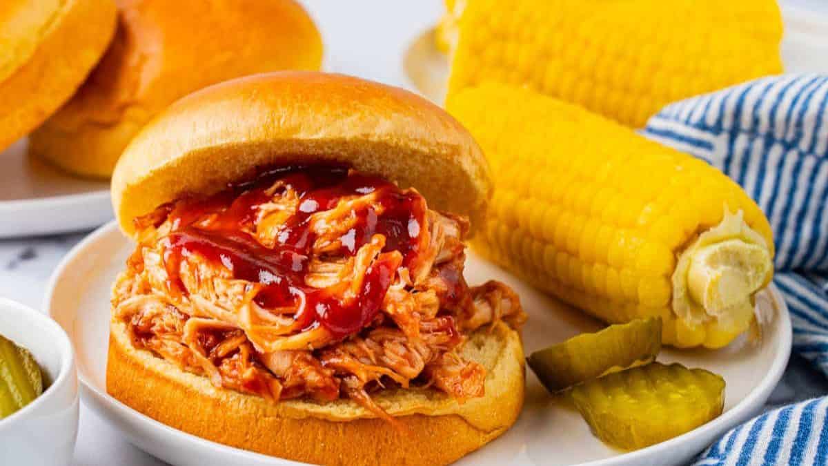 A serving of Crockpot BBQ Chicken For Pulled Chicken Sandwiches with some side corn on cob and a couple of slices of pickles. 