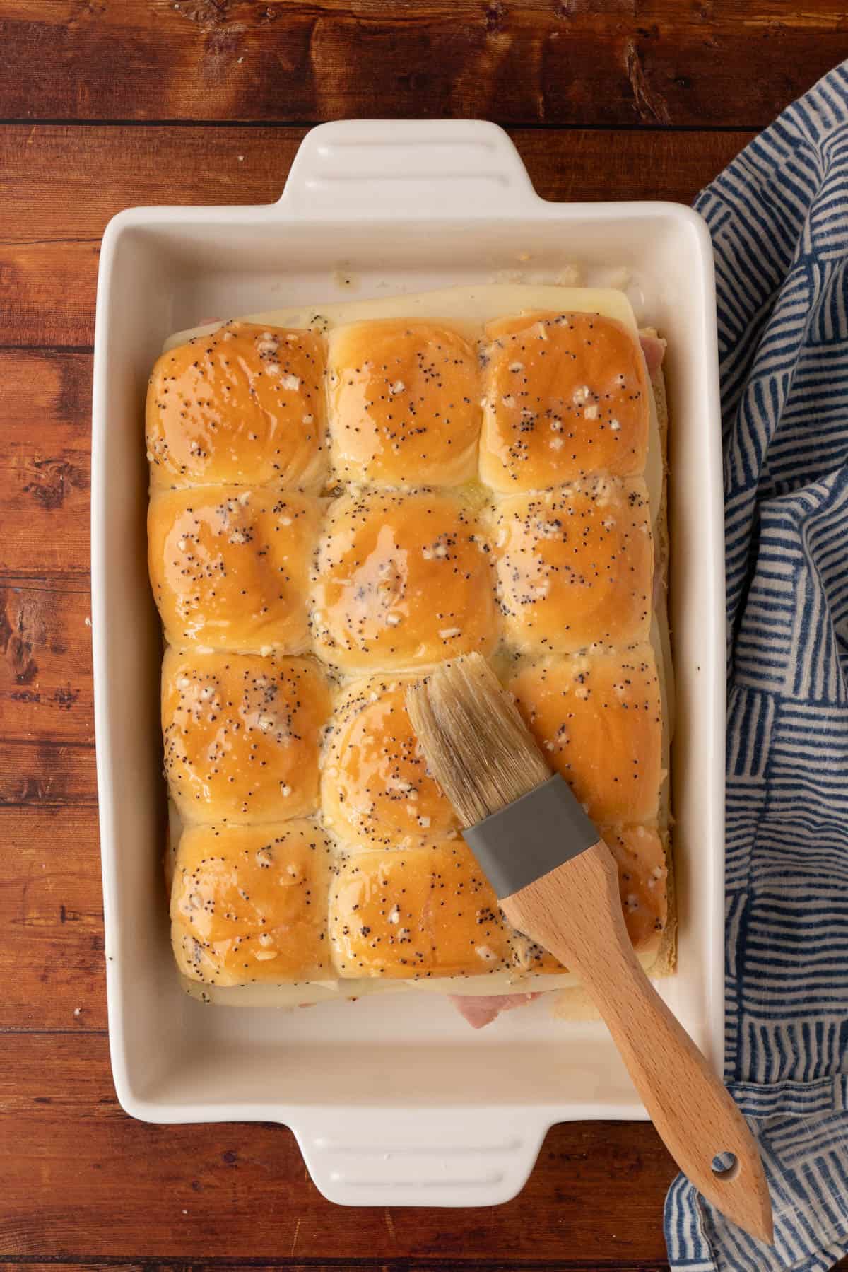 Ham And Cheese Sliders on Hawaiian Rolls on a casserole brushed with melted butter with onion, garlic, and poppy seeds and a brushed resting on the rolls.