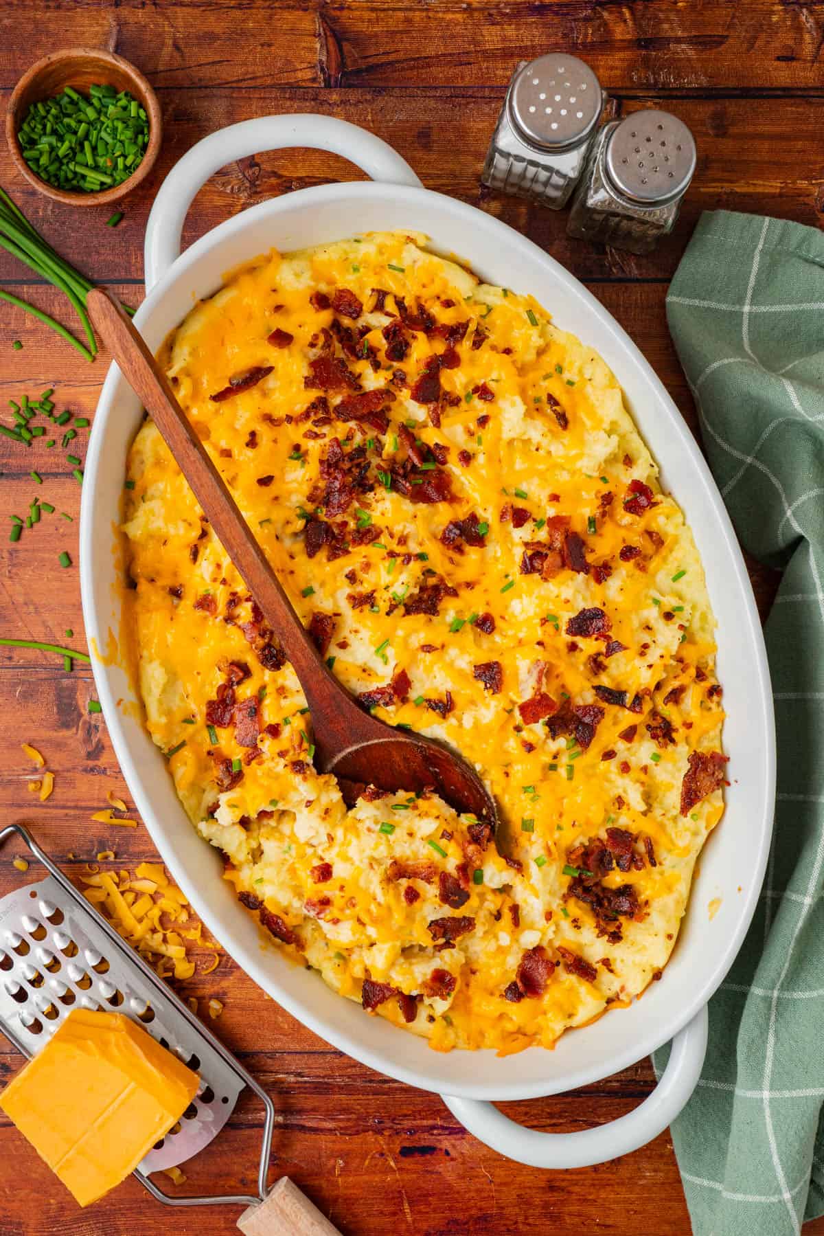 freshly cooked loaded mashed potato casserole on a wooden table with cheese, cheese grater, ever green onions, salt, and pepper bottle.