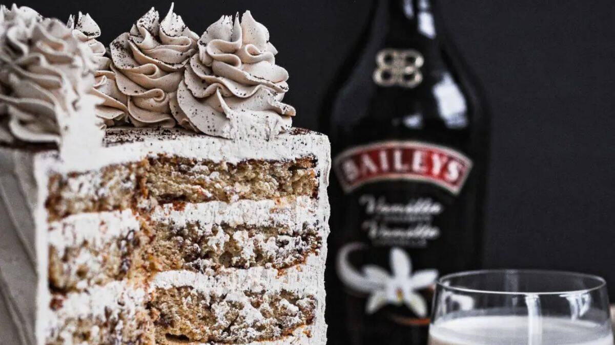 A close-up shot of Bailey's cake, a slice was taken out, which shows the layers of the cake. A blurry image of the Bailey's bottle is seen in the background.