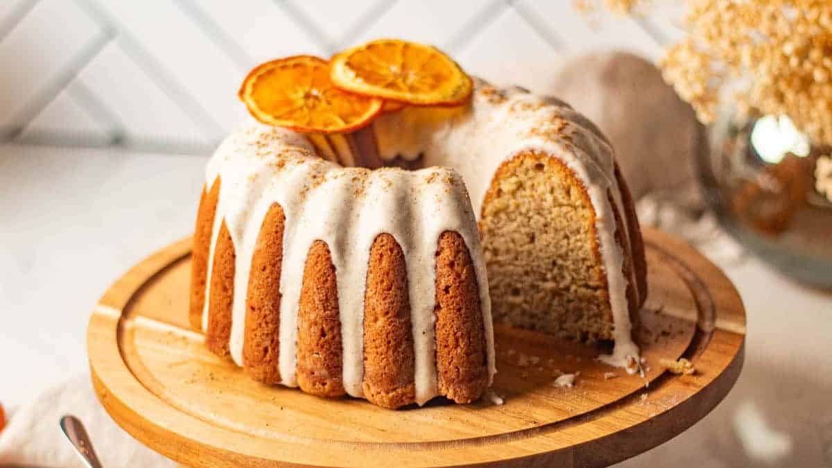 Chai Spice Bundt Cake resting on a wooden cake stand, a slice from the Bundt cake was taken that allows to have a view of the inside part of the cake, frosting is visible from the tops of the cake and two slices of orange.