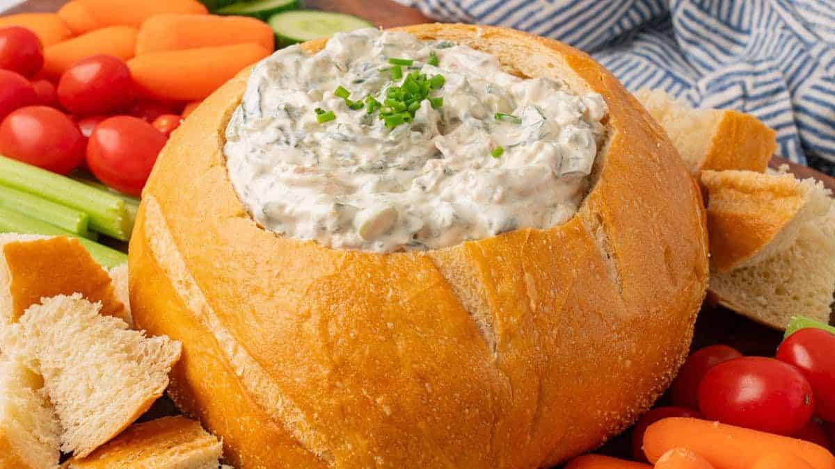 A close-up shot of Spinach dip served in a bread bowl, and some garnish on top, white surrounded with vegetables for dip and some cut of bread.