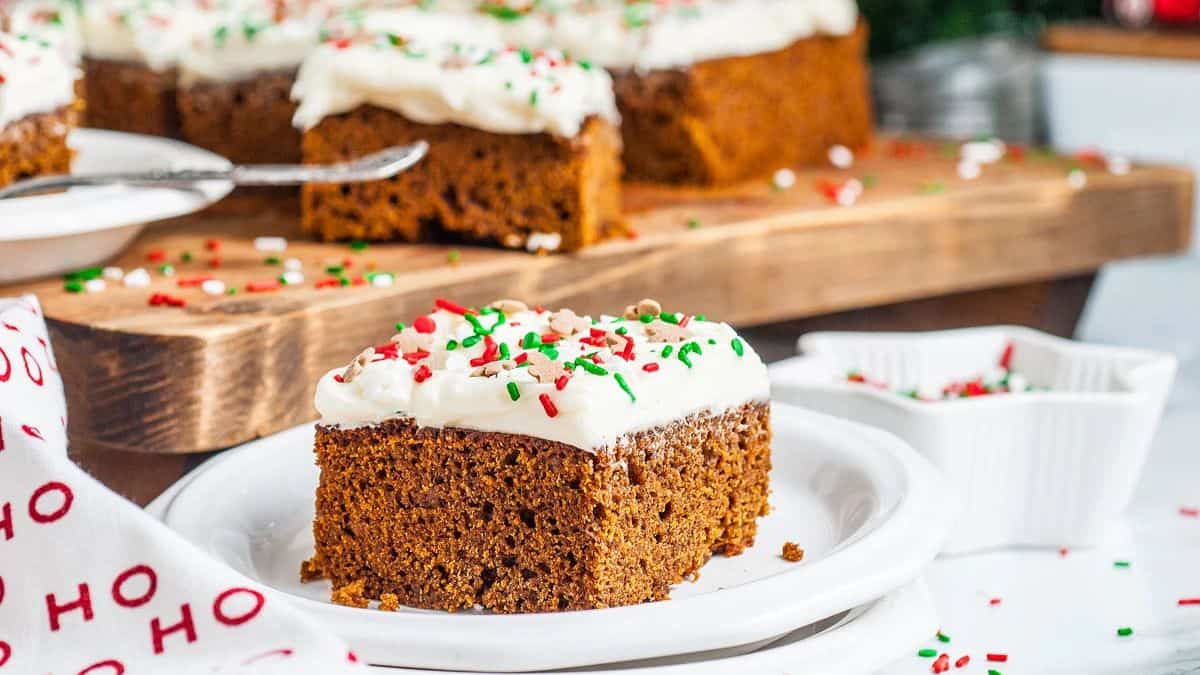 A slice of gingerbread cake served on a white plate, a blurry but visible other cake slices visible from the background as they are resting on a wooden serving board.