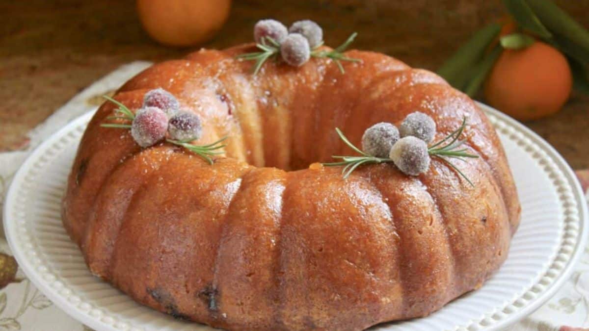 A close-up shot of a Bundt cake topped with sugared cranberries and served on a white round plate. 