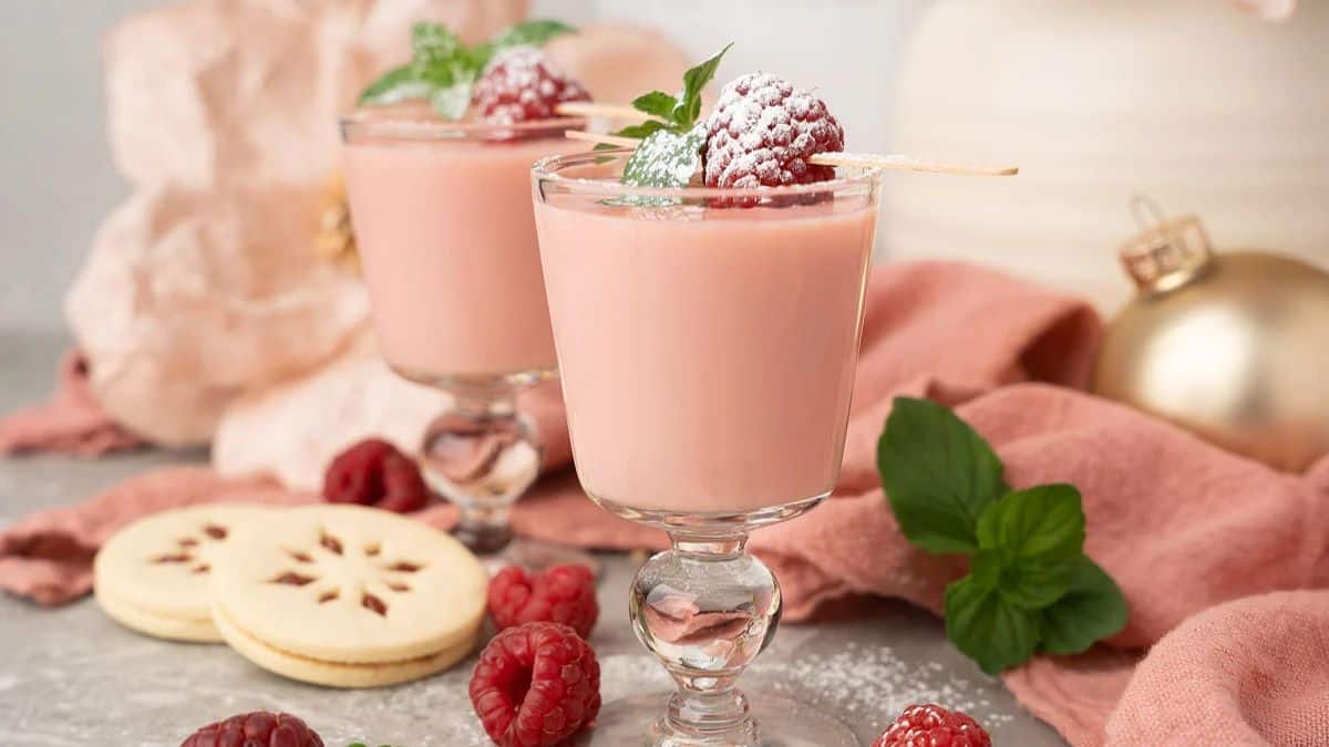 Two tall glasses of Raspberry Linzer Cookie Shots, a few raspberry cookies are found on the sides of the glasses, and a few pieces of raspberries. 