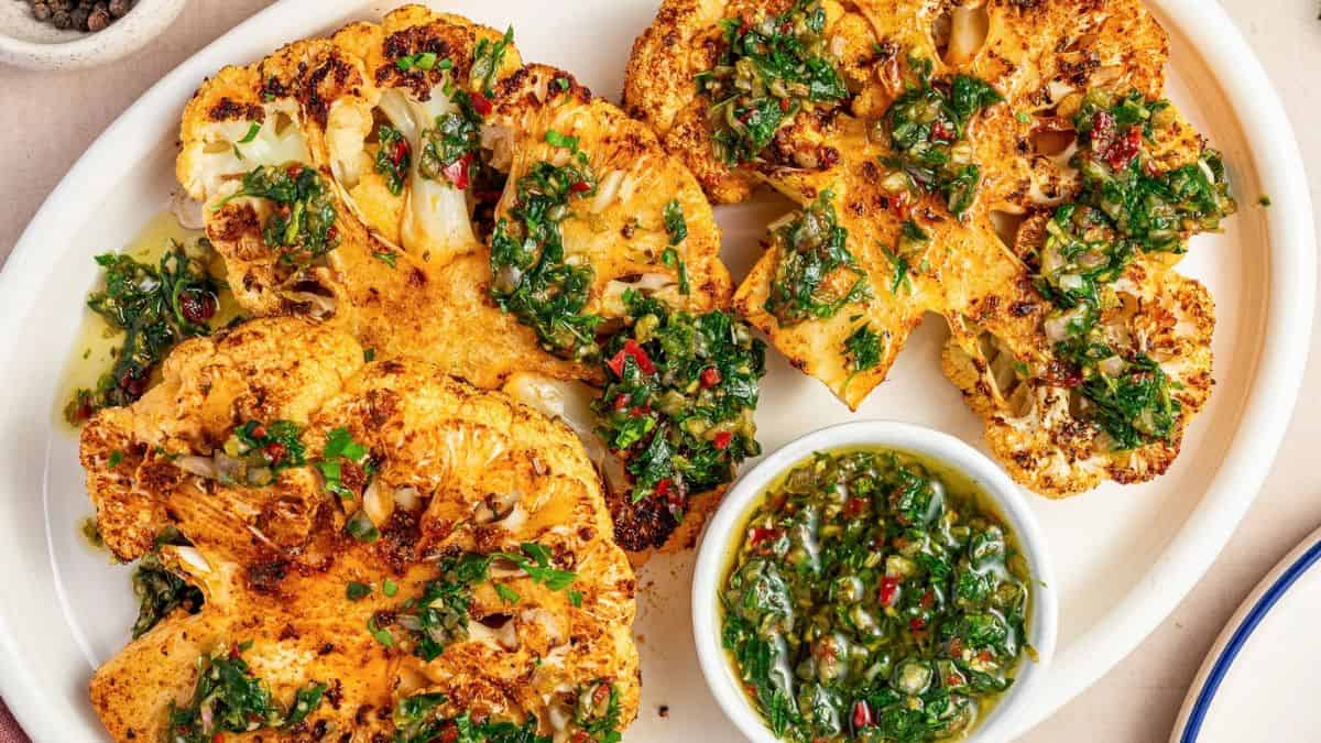 A white serving plate with three pieces of roasted cauliflower steaks and a dip on the side.