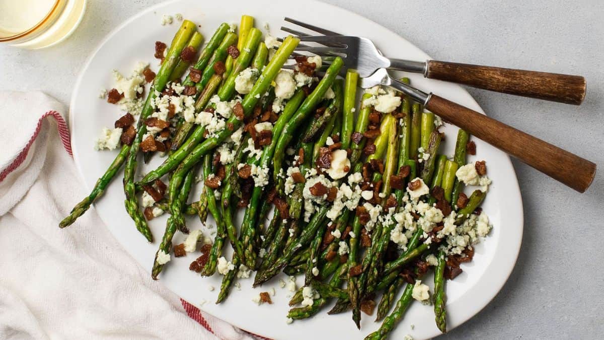 top shot of a white platter with asparagus topped with bacon bits and blue cheese pieces. 